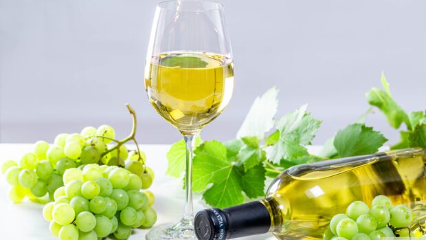A glass of white wine with green grapes and a wine bottle on a white surface