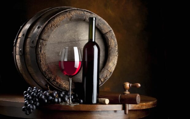 A wineglass filled with red wine next to a bottle and grapes on a wooden table