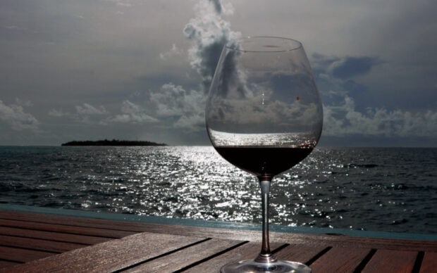 A wineglass filled with red wine reflects the ocean view and cloudy sky at sunset
