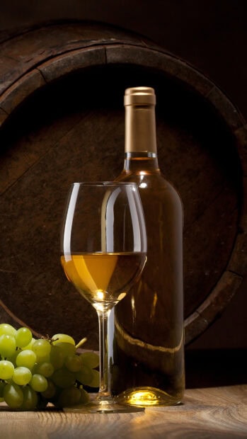 A wineglass with white wine and grapes placed in front of a wooden wine barrel