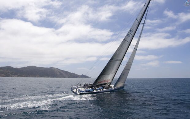 A sailing yacht cruising on open ocean near a rocky coastline under a partly cloudy sky