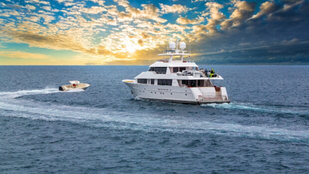 A luxury yacht cruising on the ocean water with a small boat nearby under a cloudy sky at sunset