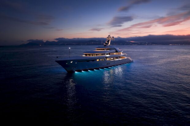 Luxury yacht illuminated at dusk on the calm sea with distant coastline visible