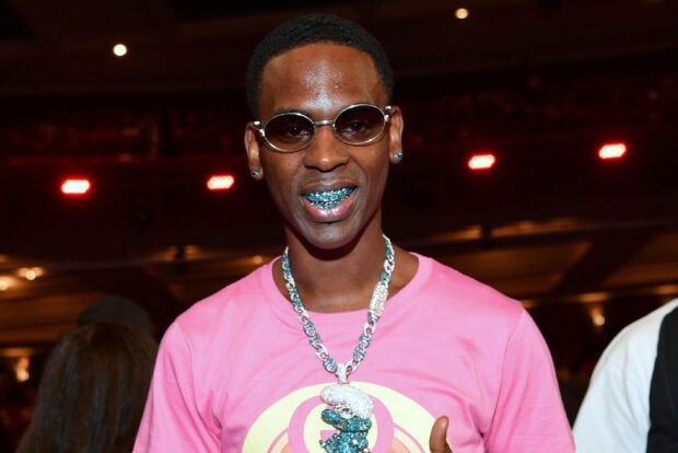 Young Dolph wearing a pink shirt and shiny jewelry smiling in a lively indoor setting