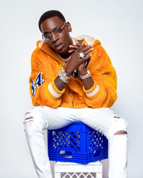 Young Dolph wearing an orange jacket and sitting on a blue crate with white ripped jeans