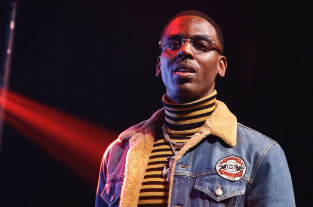 Young Dolph wearing glasses and a striped turtleneck with a denim jacket under red stage lighting