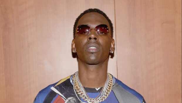 Young Dolph wearing sunglasses and multiple chains against a wooden background
