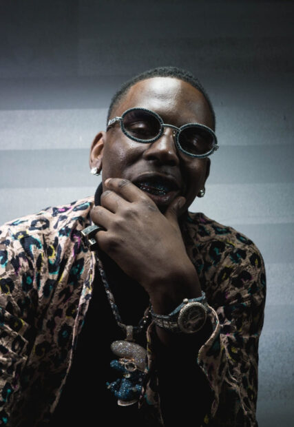Young Dolph posing with sunglasses and jewelry in a stylish patterned jacket