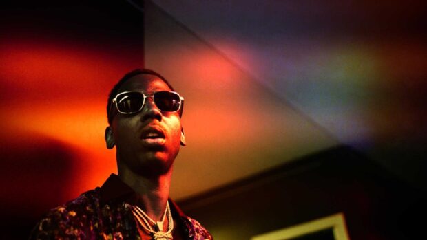 Young Dolph wearing sunglasses and chains in a dark room with colorful lighting effects