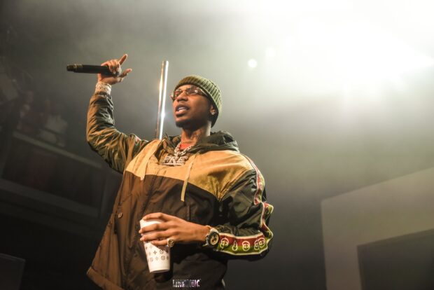Young Dolph wearing a beanie and jacket holding a microphone and cup on stage