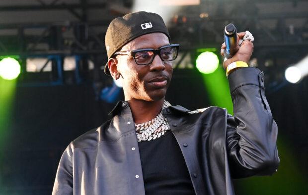 Young Dolph wearing a black leather jacket and cap while holding a microphone on stage