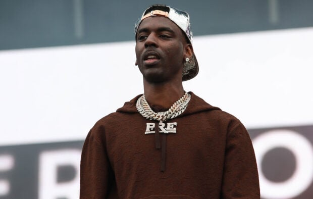 Young Dolph wearing a brown hoodie and silver chain performing on stage with a reflective cap