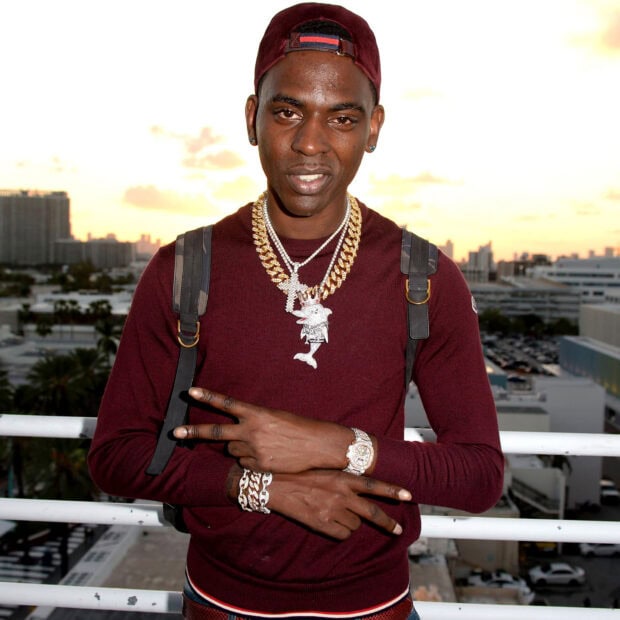 Young Dolph wearing a maroon sweater and gold chains on a city balcony at sunset