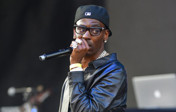 Young Dolph wearing a black leather jacket and cap holding a microphone on stage
