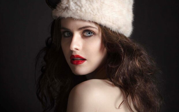 Alexandra Daddario with red lipstick and a white furry hat looking over her shoulder