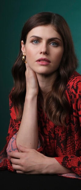 Alexandra Daddario in a red patterned dress posing with her hand on her neck against a green background