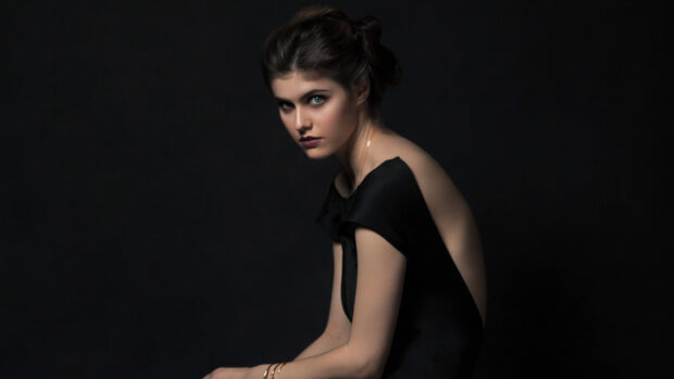 Alexandra Daddario wearing a black dress with an open back sitting against a dark background