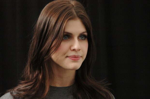 Alexandra Daddario with long brown hair and blue eyes looking thoughtfully against a dark background