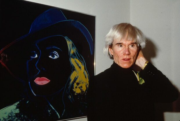 Andy Warhol standing beside a colorful portrait artwork in a gallery setting