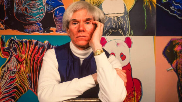 Andy Warhol dressed in white and blue sitting in front of colorful animal artwork