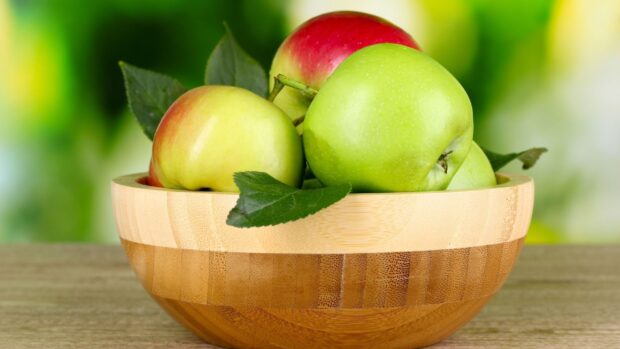 A wooden bowl filled with fresh apple fruit and green leaves on a wooden surface