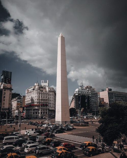 The Argentina obelisk stands tall in the city center with busy traffic around including taxis and pedestrians