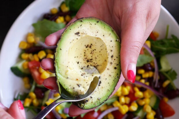 A hand holding an avocado half with a spoon scooping avocado flesh sprinkled with black pepper