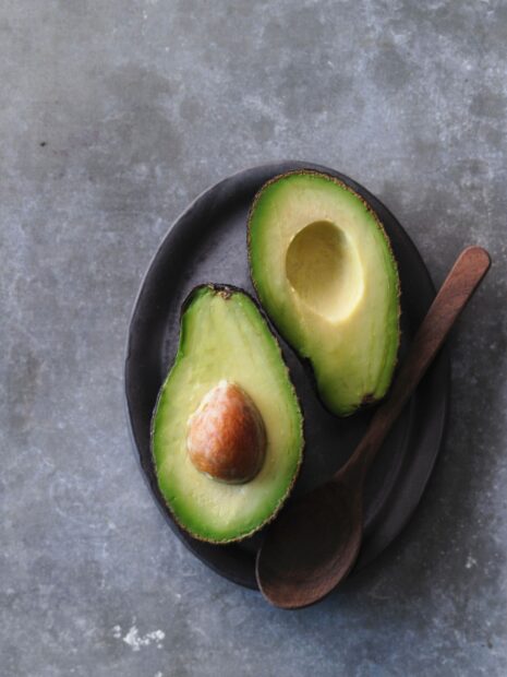 Fresh avocado halves with seed on a dark plate next to a wooden spoon on a gray surface