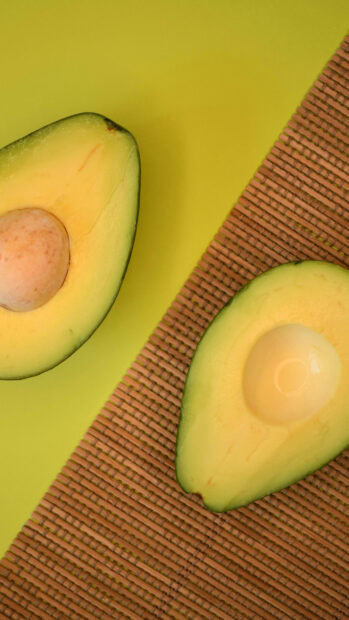 Two halves of fresh avocado showing seed and flesh on a woven mat and yellow surface