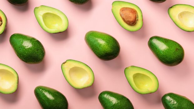 Fresh avocado halves and whole avocados arranged on a pink surface in a repeating pattern