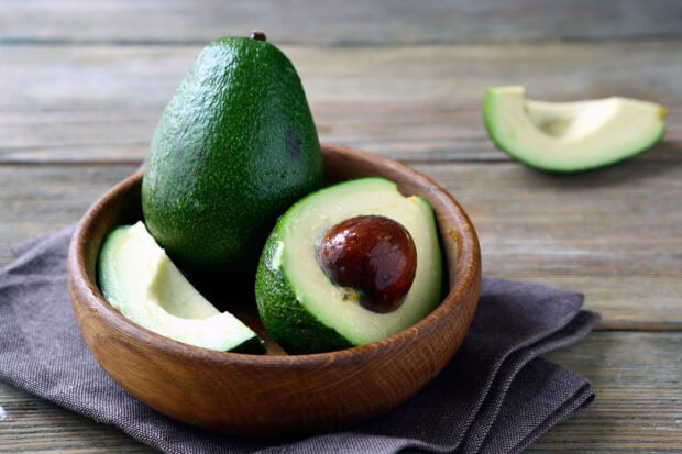 Fresh avocado halves with seed and slices in a wooden bowl on a rustic table