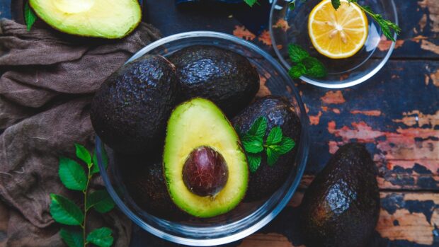 Fresh avocado fruit with pit in a bowl surrounded by whole avocados and lemon slice on wooden surface
