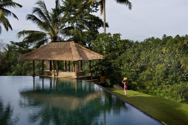 A traditional hut near a tropical forest in Bali showing lush greenery and a person carrying a basket on their head