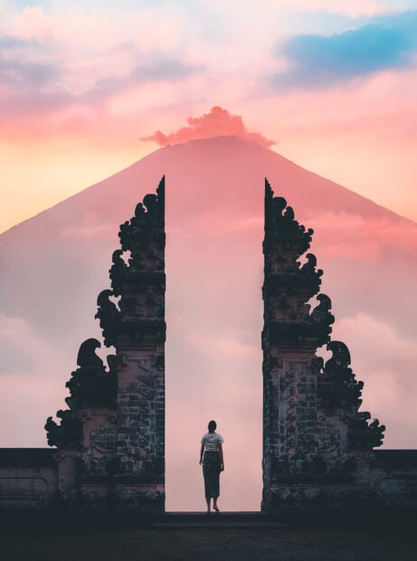 Woman standing at a traditional gate with Bali volcano in the background during sunset