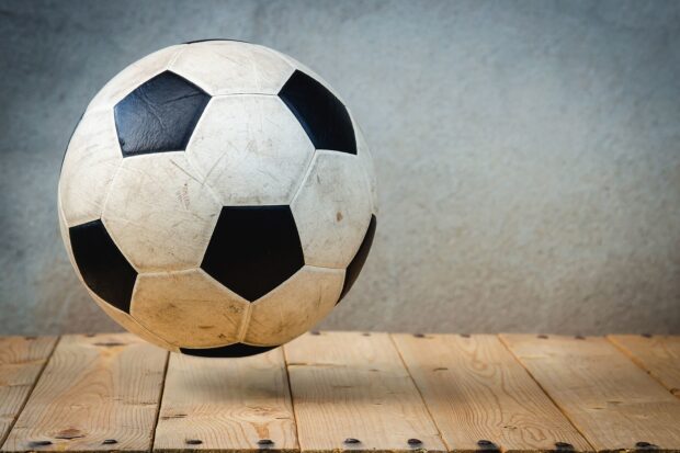 A worn soccer ball hovering above a wooden surface with a plain gray wall in the background
