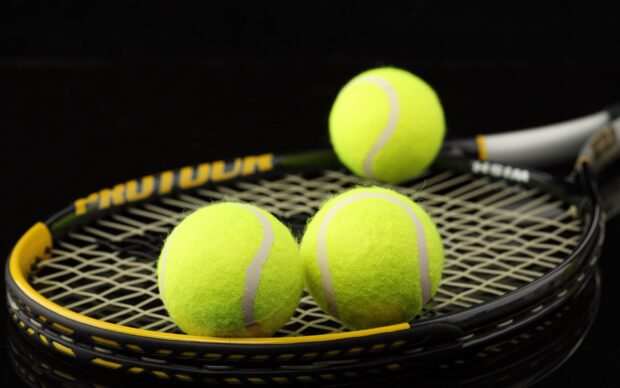 Three tennis balls resting on a black tennis racket with yellow accents on a dark background
