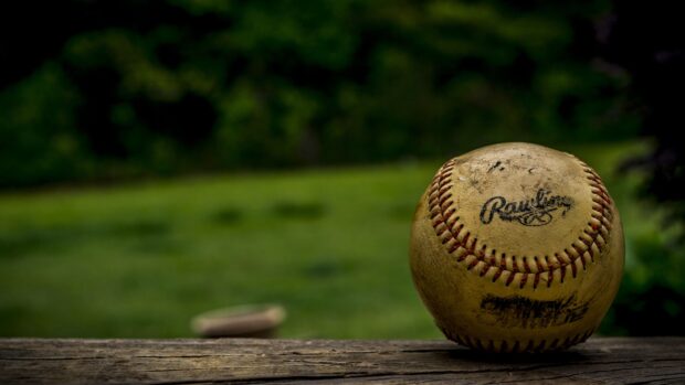 A close up of a baseball resting on a wooden surface outdoors with green grass in the background