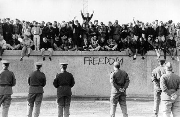 People gathered on the Berlin Wall with freedom graffiti and soldiers standing below