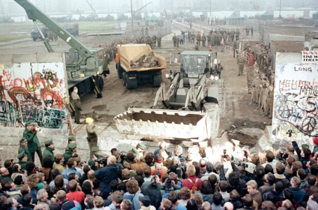 A crowd watches military vehicles dismantle the Berlin Wall on a historic day