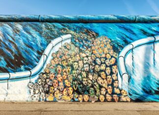 A colorful mural on the Berlin Wall showing many expressive faces merging together under a blue sky