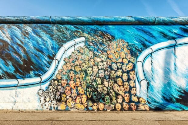 A colorful mural on the Berlin Wall showing many expressive faces merging together under a blue sky