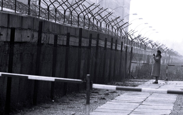 A woman standing near the Berlin Wall with barbed wire and fences surrounding the area