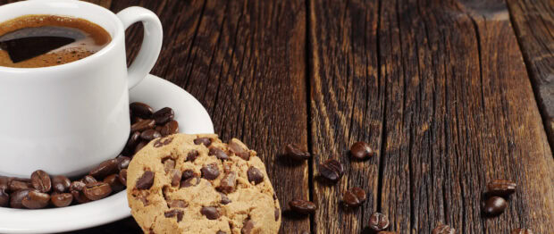 A chocolate chip biscuit placed on a wooden table next to coffee beans and a white cup of coffee