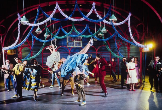 A lively Broadway dance scene featuring performers in colorful costumes and dynamic choreography