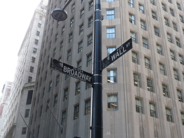 Street signs showing Broadway and Wall street in a cityscape with tall buildings