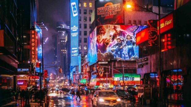 Nighttime street scene in Broadway with neon signs and Stranger Things billboard on a rainy evening