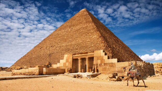 Ancient pyramid at Cairo Egypt with a man riding a camel nearby under a blue sky