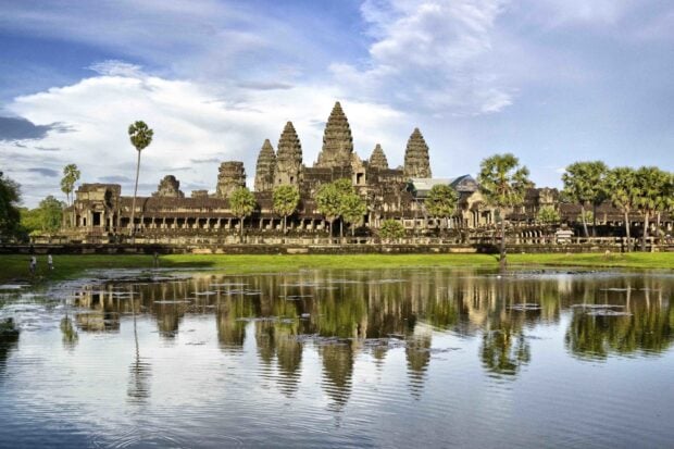 Ancient Cambodia temple reflecting on water at sunset with palm trees and cloudy sky