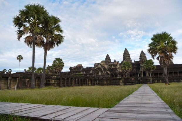 Ancient Cambodia temple surrounded by palm trees and grassy field under a cloudy sky