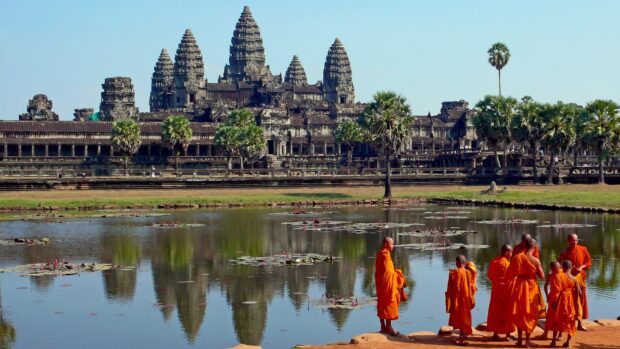 Ancient Cambodia temple reflected in water with monks wearing bright orange robes standing nearby